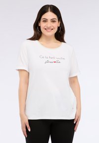 Camiseta blanca de manga corta con cuello redondo, que presenta texto impreso en negro y un acento en forma de corazón rojo. Fabricada con tejido suave y con un corte relajado.