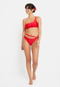 Raudona bikini maudymosi kostiumėlis su vieno peties viršumi ir kryžminėmis juostomis apačioje, turintis apvalų detalių elementą ir lygų medžiagos tekstūrą.