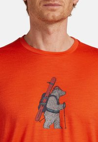 Oranžové tričko s krátkým rukávem, které zepředu zdobí detailní grafika medvěda nesoucího lyže a vybavení, obrysovaná černě.