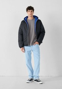 Giacca a vento nera con fodera interna blu, t-shirt grigia, jeans azzurri chiari e sneaker nere. Modello in piedi contro uno sfondo bianco.