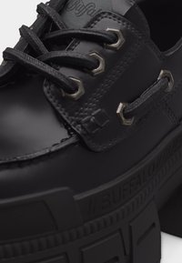 Μαύρο δερμάτινο παπούτσι Buffalo με χοντρή σόλα, στρογγυλές μεταλλικές οπές και μαύρα κορδόνια, που παρουσιάζει λεπτομερείς ραφές και ανάγλυφο το όνομα της μάρκας στη σόλα.