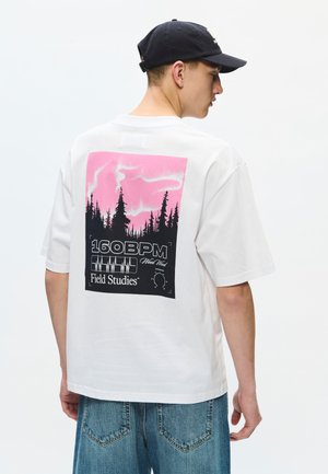 Jeune homme portant un t-shirt blanc avec un motif de forêt rose et noir et le texte "160 BPM Field Studies" au dos.