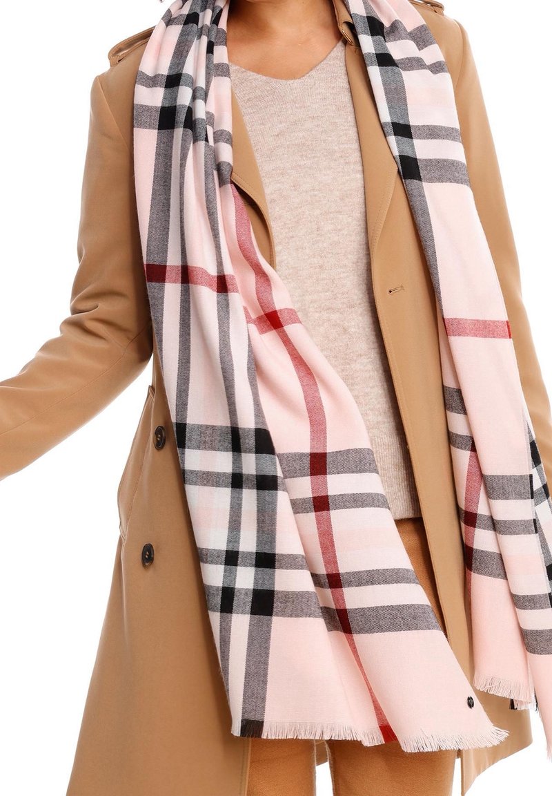 Femme portant un trench beige, un pull beige clair et une grande écharpe à carreaux rose avec des rayures noires, grises et rouges posée sur les épaules.
