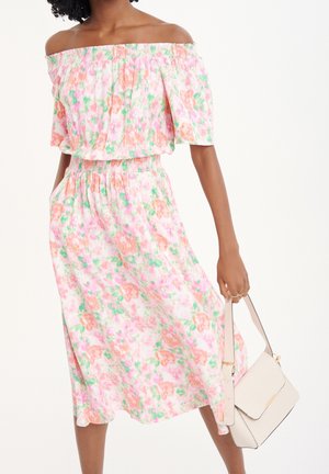 Donna che indossa un vestito rosa floreale scollato sulle spalle e tiene una borsa bianca con tracolla su uno sfondo neutro.