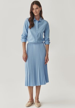 Blauwe overhemd met opgerolde mouwen en een geplisseerde midi-rok, beide in lichtblauw, gecombineerd met beige platte schoenen.
