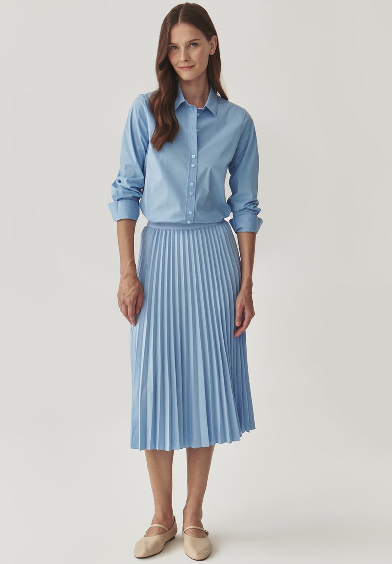 Blauwe overhemd met opgerolde mouwen en een geplisseerde midi-rok, beide in lichtblauw, gecombineerd met beige platte schoenen.