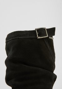 Botte de cheville en suède noir avec un revers et un détail de boucle en métal argenté. Texture lisse avec des coutures visibles le long des bords.