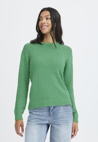 Mujer con cabello largo y oscuro que lleva un suéter verde texturizado y jeans azul claro, sonriendo y mirando de lado frente a un fondo blanco.