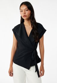 Zwarte wikkeltop met brede kraag, korte mouwen en een strik in de taille. Heeft een textuurstof en een diepe V-hals. Gekleed met witte broeken.