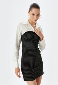 Schwarz-weiß gestreiftes Kleid mit tailliertem Oberteil und ausgestellt Rock, kombiniert mit einem weißen Hemd mit Kragen. Strukturierter Stoff mit vertikalen Linien.