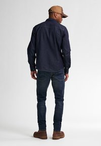Camisa de mezclilla azul oscuro de manga larga, con detalle en el yugo trasero, combinada con jeans ajustados azul oscuro y botas de tobillo marrones. Fondo claro.