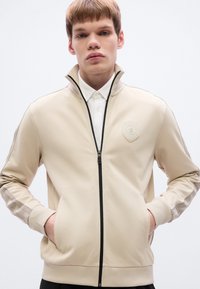 Μπεζ μπουφάν με φερμουάρ από απαλό ύφασμα, με ριμπ μανσέτες, πλαϊνές τσέπες και υφασμάτινο έμβλημα στο στήθος, με μαύρες λεπτομέρειες.