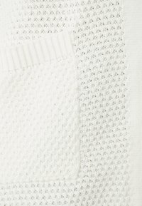 Pull blanc en maille présentant des textures variées, dont un motif alvéolé et des accents côtelés, avec une poche plaquée à l'avant.