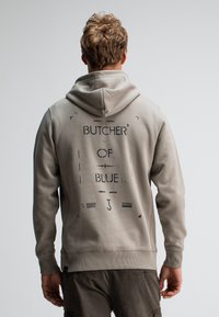 Lichtgrijze hoodie met een zwart grafisch ontwerp waarop "BUTCHER OF BLUE" op de achterkant staat. Het materiaal lijkt zacht, met ribbels aan de mouwen en onderkant.