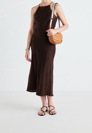 Femme portant une robe midi marron sans manches, des sandales marron à brides et un petit sac bandoulière beige avec une sangle blanche à logo.