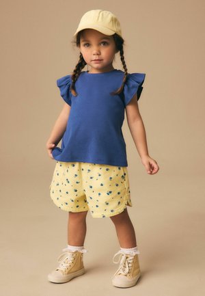 Jong meisje met gevlochten haar, een gele pet, een blauw topje met ruches zonder mouwen, gele korte broek met bloemenprint, witte sokken en beige sportschoenen.