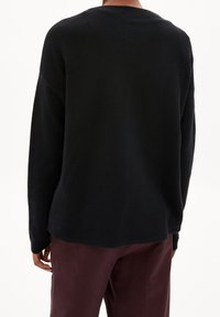 Osoba v černém volném pleteném svetru s dlouhými rukávy a tmavě vínových kalhotách, viděná zezadu na jednobarevném bílém pozadí.