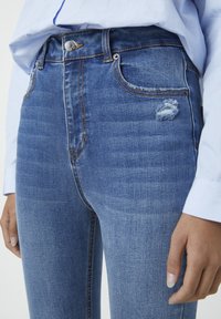 Jeans de tiro alto en denim azul de grosor medio con un ligero estiramiento, que presentan un bolsillo desgastado, costuras en contraste y un cierre de botón plateado.