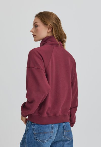 Bordo pulover z visokim ovratnikom, sproščenega kroja, rebrastimi manšetam in robom. Mehka tekstura, vidni šivi in minimalističen dizajn.