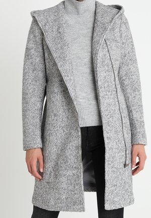 Long manteau gris texturé avec capuche porté sur un pull à col roulé côtelé gris clair et un pantalon noir.