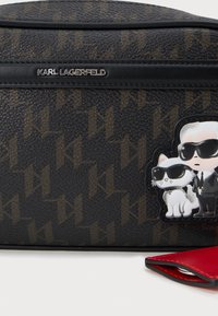 Melnā teksturētā soma ar brūnu logo rakstu. Tam ir karikatūras uzlīme ar vīrieti un kaķi saulesbrillēs. Sarkanā akcenta detaļa uz rāvējslēdzēja.