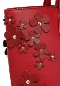 Červená taška typu tote z umělé kůže ozdobená několika texturovanými květinovými aplikacemi a zlatými cvočky, s pevnou strukturou a dvojitými rukojeťmi.