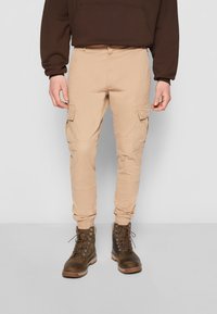 Pantalon cargo beige avec poignets élastiques, poches latérales et devant plat, associé à des bottes marron à lacets. Design décontracté pour une tenue extérieure.