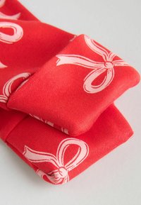 Tessuto rosso con una consistenza morbida, caratterizzato da un motivo ripetuto di fiocchi di nastro bianchi. I polsini sono piegati, mostrando una finitura liscia e un colore vivace.