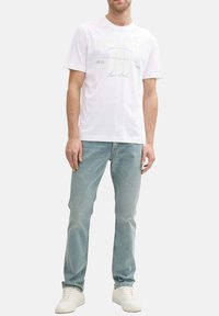 T-shirt en coton blanc avec des graphismes textuels en gris clair et des accents jaunes, associé à un jean bleu clair et des baskets blanches.