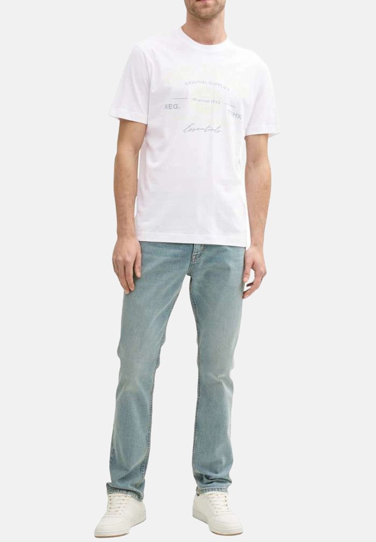 T-shirt en coton blanc avec des graphismes textuels en gris clair et des accents jaunes, associé à un jean bleu clair et des baskets blanches.