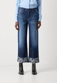 Široke denim hlače temno modre barve z vezenimi cvetličnimi vzorci in razfrakanim robom. Ujemajo se z črnimi gležnjarji.
