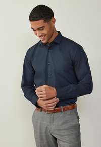 Dunkelblauses Langarmhemd mit spitzem Kragen und Knopfverschluss, kombiniert mit grauen karierten Hosen und einem braunen Leder-Gürtel.