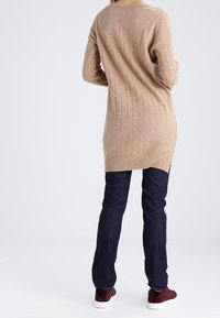 Personne portant un long pull beige côtelé, un jean bleu foncé et des baskets bordeaux, debout, dos à la caméra.