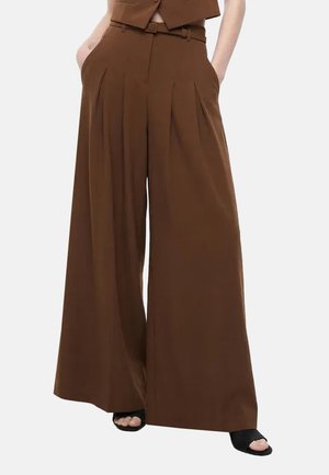 Pantaloni marroni a vita alta e gamba larga con pieghe anteriori, indossati con tacchi neri open toe, mani nelle tasche e un gilet marrone coordinato.