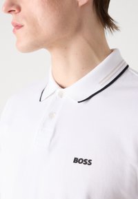 Biele polo tričko s čiernymi akcentami a logom. Obsahuje klasický golier, gombíkovú légu a tri vodorovné pruhy na golieri.