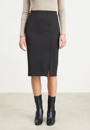 Kobieta ubrana w czarną ołówkową spódnicę do kolan z rozcięciem z przodu, beżową bluzkę z długim rękawem i czarne botki, stojąca na neutralnej podłodze.