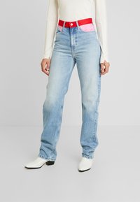 Kvinna som bär ljusblå jeans med röd midja och rosa fickfoder, vit långärmad tröja och vita klackstövlar, stående på en enfärgad bakgrund.