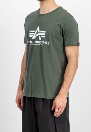 Man draagt een olijfgroen Alpha Industries t-shirt met wit logo en tekst, gecombineerd met losse zwarte broek, staand tegen een witte achtergrond.