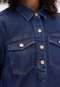 Gros plan sur une chemise en denim bleu foncé avec col, poches poitrine et boutons en cuivre gravés.