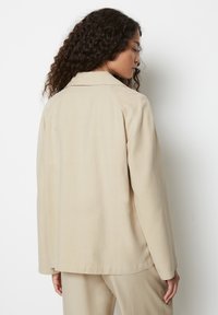 Beige jakk, millel on lõtv istuvus, pehme tekstuur, sälguga krae ja pikad varrukad, shown tagantpoolt, minimaalse detailitusega.