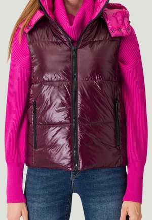 Femme portant un gilet matelassé brillant couleur bordeaux sur un pull côtelé rose vif et un jean bleu, debout les bras détendus.