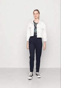 Jaqueta de ganga branca cortada, blusa floral preta, calças azul-marinho e sapatilhas pretas. Modelo de pé com um fundo neutro.