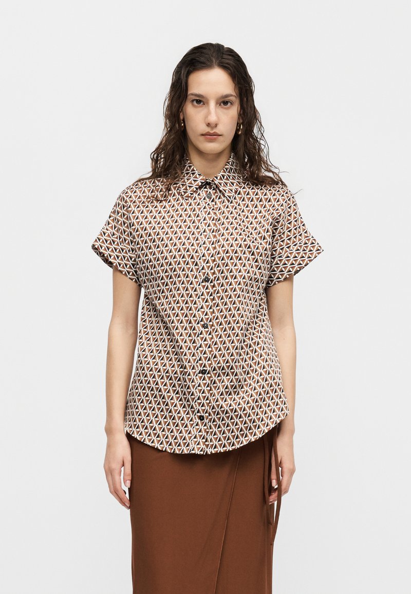 Γυναίκα που φοράει καφέ πουκάμισο με γεωμετρικό μοτίβο και κοντά μανίκια και καφέ φούστα, στέκεται μπροστά σε απλό φόντο.