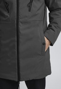 Veste rembourrée grise avec fermeture éclair à l'avant, cordons de serrage réglables et poches latérales. Texture lisse et design simple.