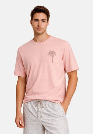 Ung mand iført en lyserød t-shirt med palmetræsgrafik og lysegrå mønstrede shorts med snøretræk, står med den ene hånd i lommen.