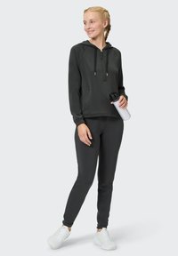 Dunkelgrauer Hoodie mit halbseitigem Reißverschluss und Fronttasche, kombiniert mit passenden Jogginghosen. Das Model hält eine weiße Wasserflasche und trägt weiße Sneakers.