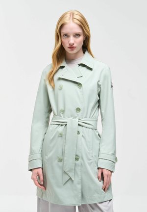 Jonge vrouw met lang blond haar, gekleed in een lichtgroene trenchcoat met ceintuur, staand tegen een effen lichte achtergrond.