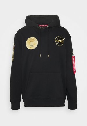 Sort NASA-hoodie med gyldne missionsmærker, front-kængurulomme, sorte snore og rød "Remove Before Flight"-mærkat på lynlåslommen på venstre ærme.