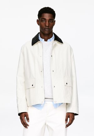 Jasnobeżowa kurtka z czarnym kołnierzem ze sztruksu, zapinana na zatrzaski, z bocznymi kieszeniami. Noszona na szarym swetrze i jasnoniebieskiej koszuli.