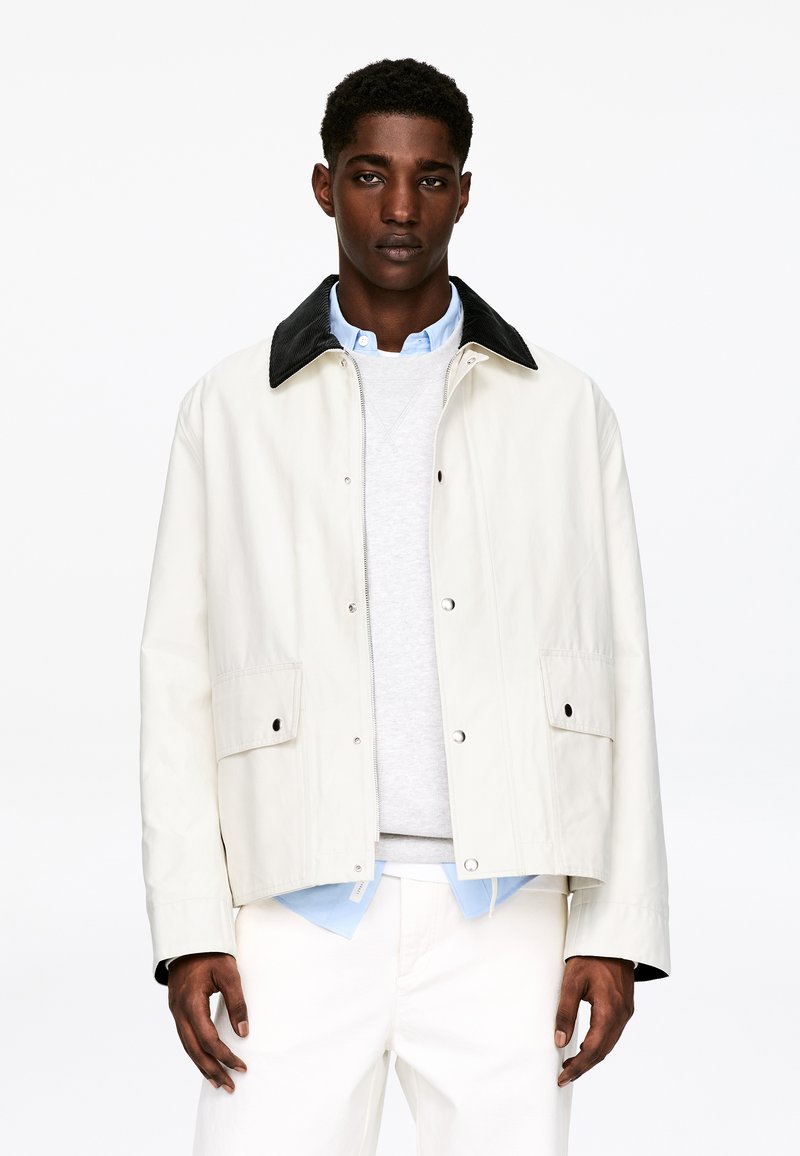 Veste beige clair avec un col en velours côtelé noir, dotée de fermetures à pression et de poches latérales. Portée sur un pull gris et une chemise bleu clair.